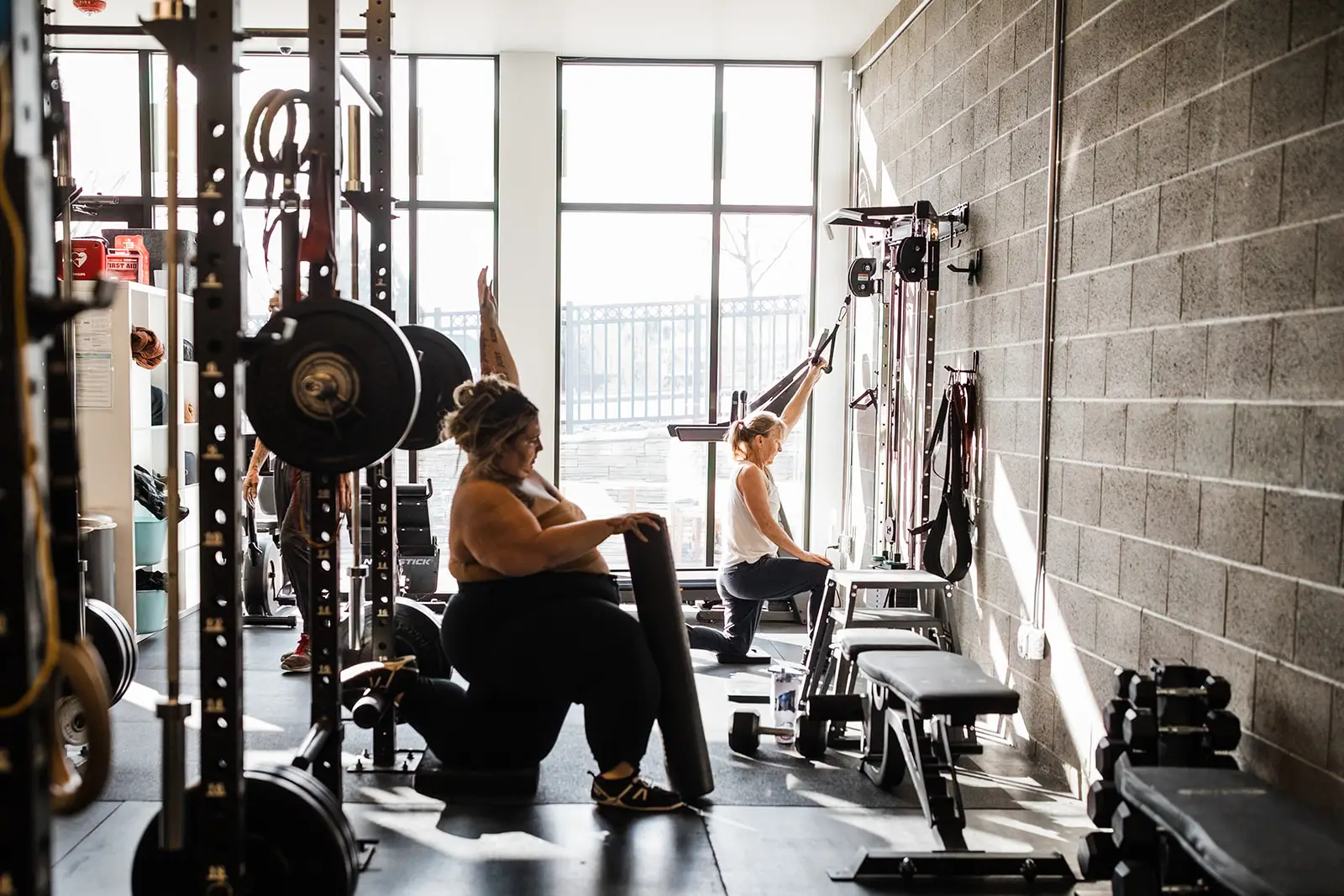 A woman performing a hip flexor stretch at a squat rack in the foreground, and second woman performing a lat pulldown in the background