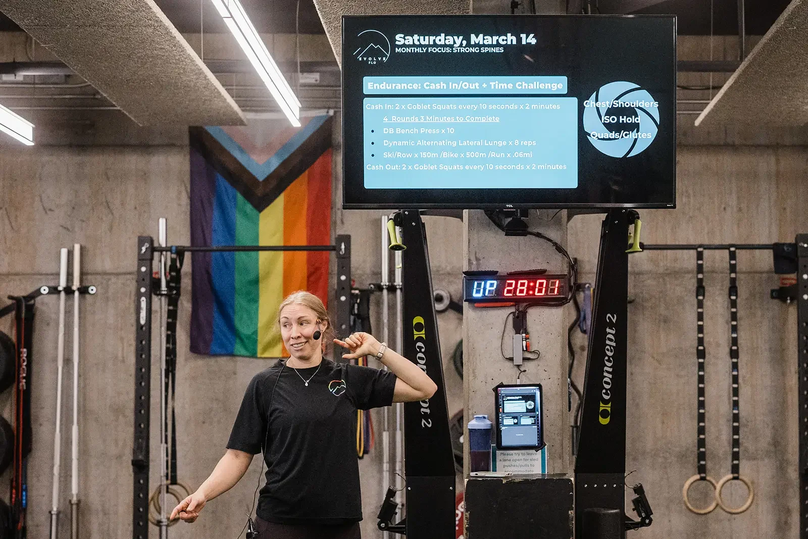 Female strength coach teaching and smiling underneath a TV that displays the strength workout