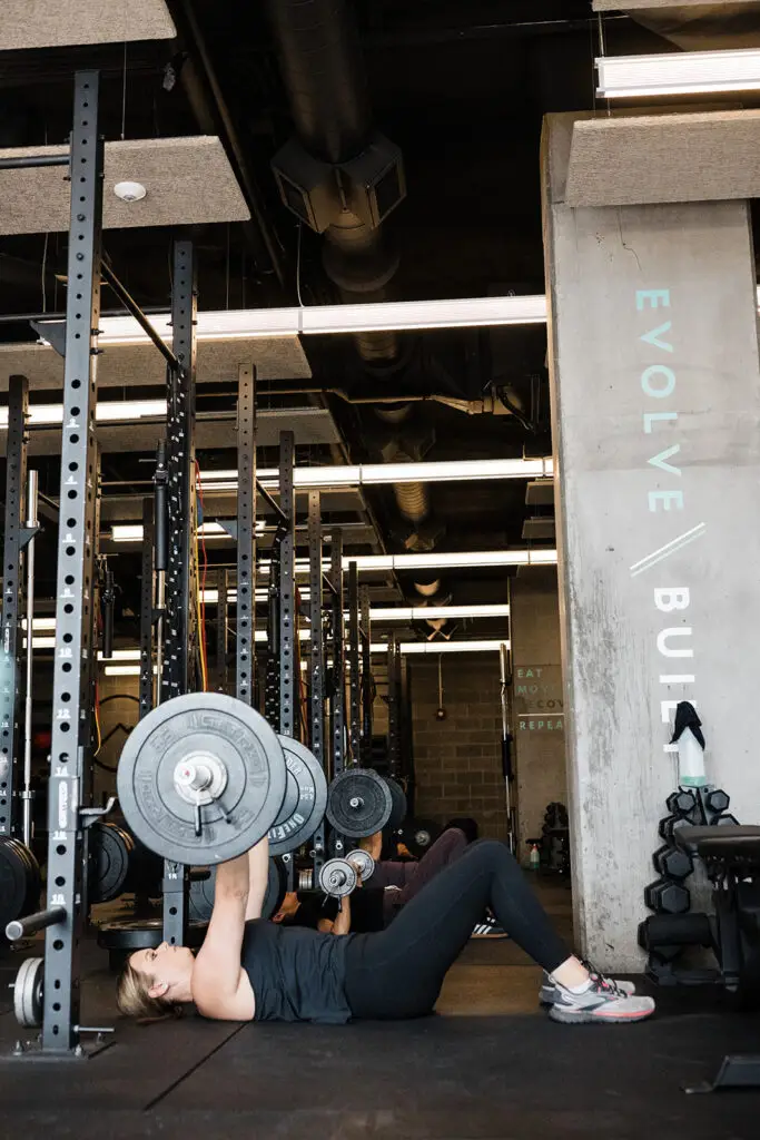 Image of a woman performing a barbell floor press next to a concrete column that with the word EVOLVE Built vertically