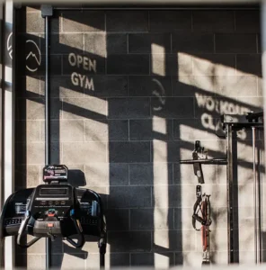 Warm sunset light streams through a gym window, casting the words 'Open Gym' and 'Workout Classes' across a cinderblock wall above a treadmill.